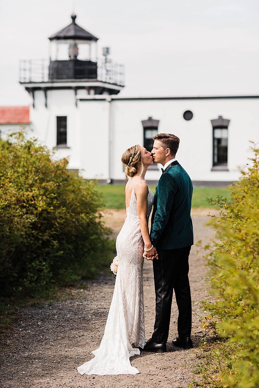 A bride kisses her groom, wearing a green suit, in front of a lighthouse in Washington.