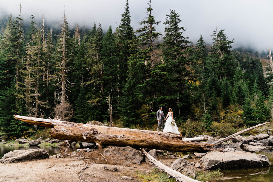 A bride and groom walk onto a large log in the Washington backcountry on their elopement day