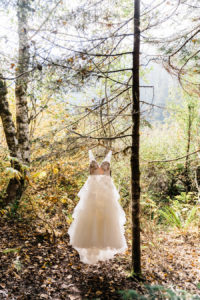 A layered wedding dress hangs from a tree in the woods backlit by the sun coming through the trees