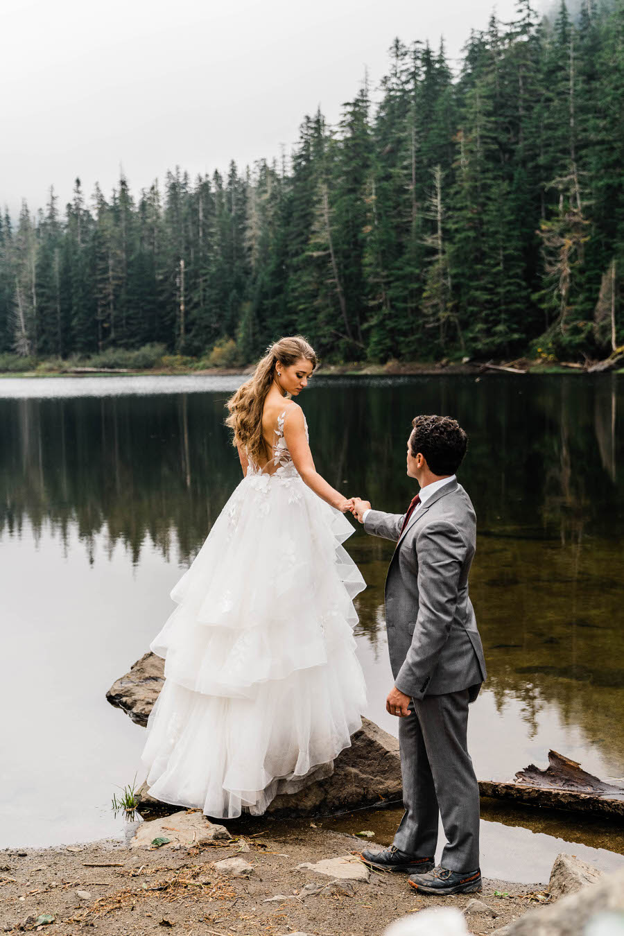 A bride stands on a rock and looks back at her groom next to an alpine lake