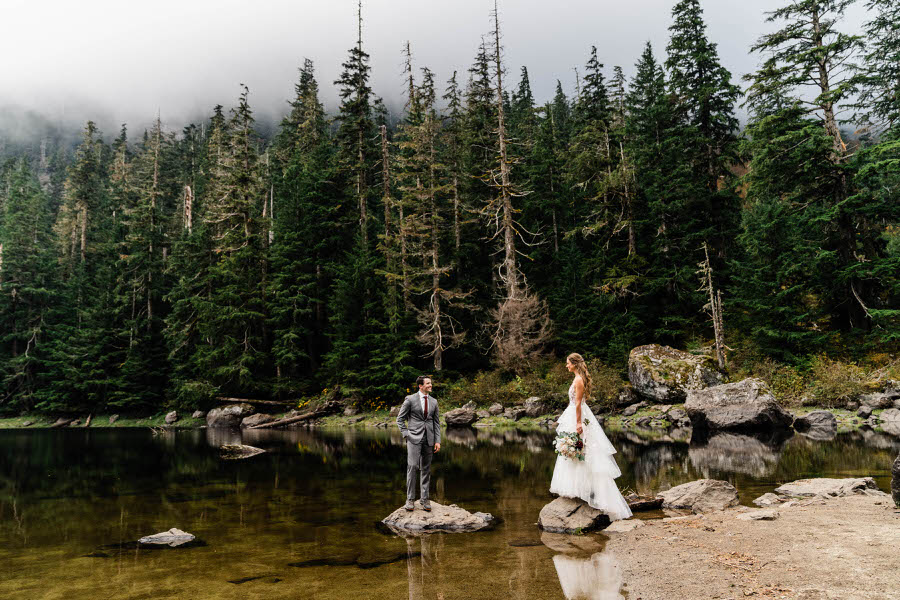A bride and groom stand on small rocks in an alpine lake in the Washington backcountry
