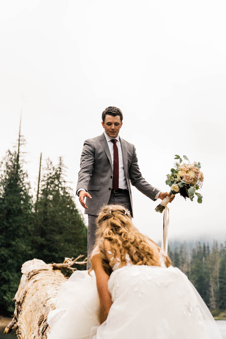 A groom reaches down for his bride's hand as they climb up a giant fallen log