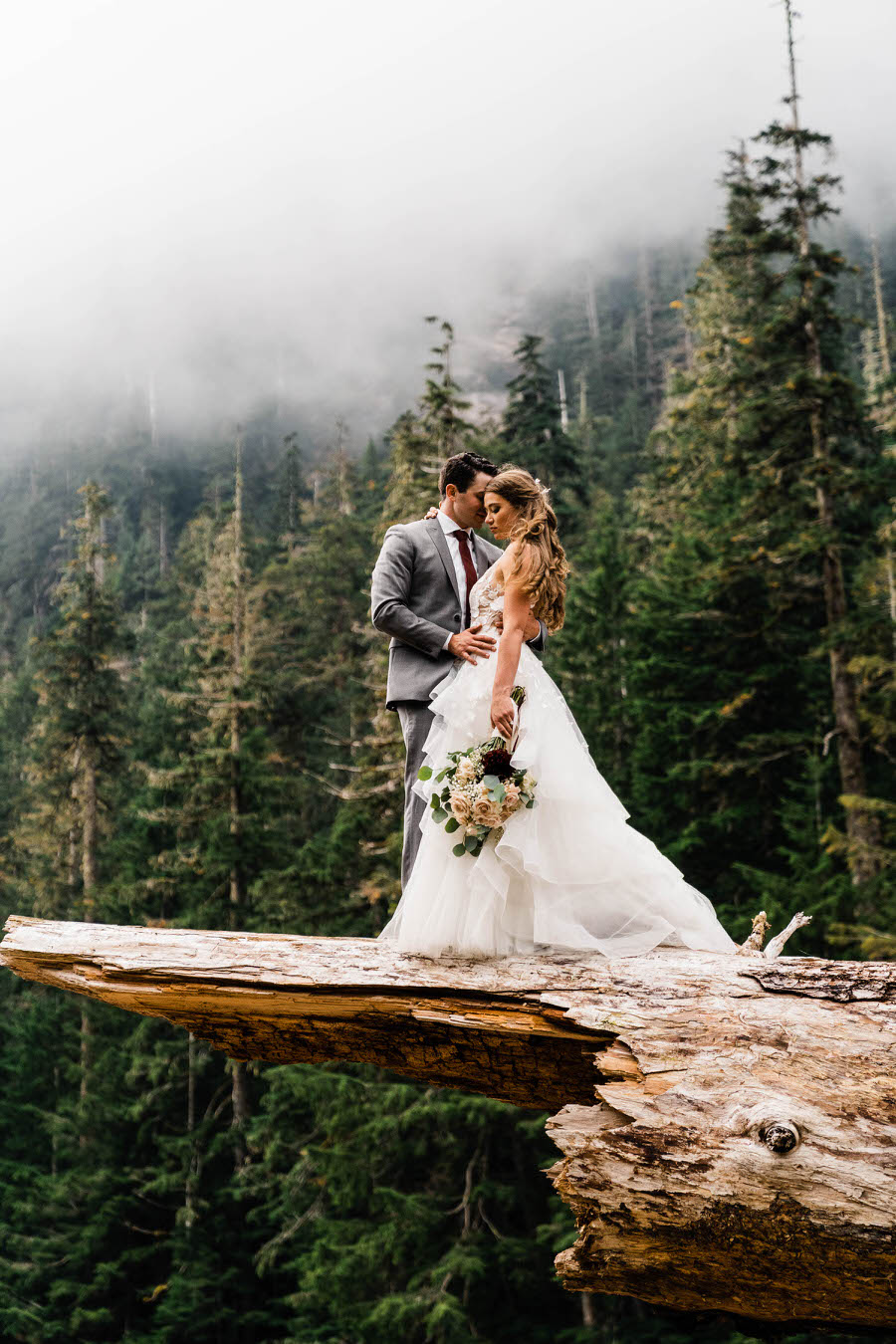 A bride and groom stand on a giant fallen log in the Washington backcountry after their mountain wedding in the backcountry