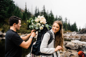 A groom helps his bride pack her bouquet in her backpack for their hiking elopement in the Washington cascades