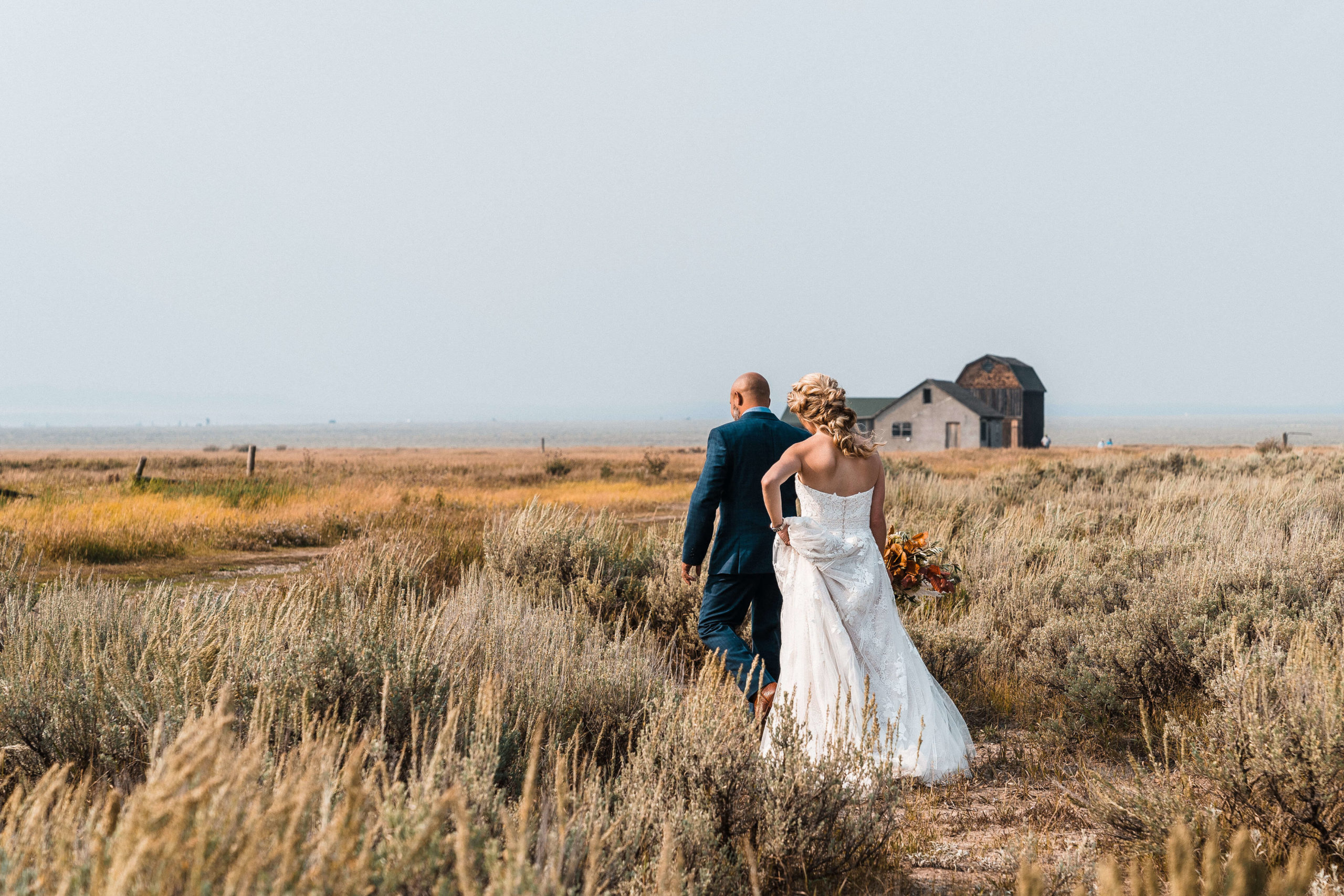 Bride and groom walking through a field towards a barn