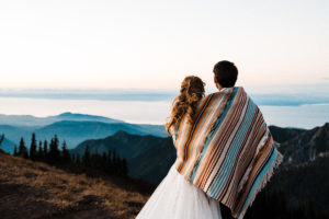 Bride and groom wrapped in blanket staring out at the sunset over the mountains