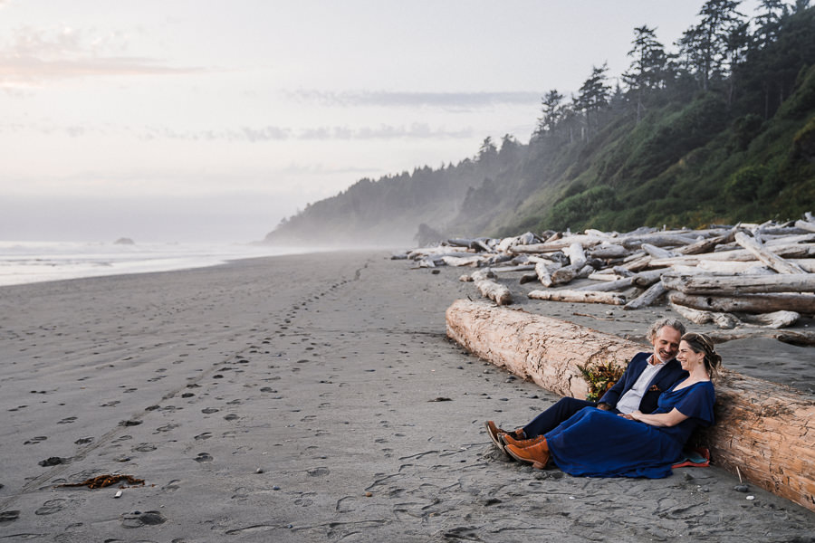 Bride and groom lean against a log and laugh together on Kalaloch Beach after their elopement ceremony