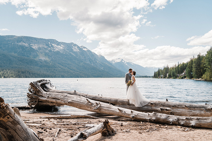 a bride and groom stand on a log at lake wenatchee just outside of leavenworth