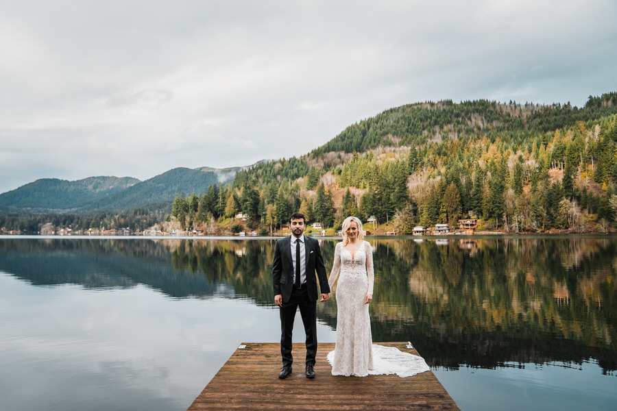 A bride and groom hold hands while standing on a dock with mountain views on Lake Sutherland
