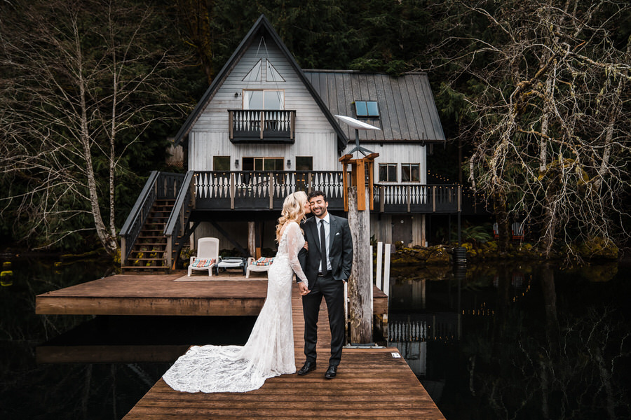 A bride kisses her groom on the cheek in front of a lake cabin on Lake Sutherland