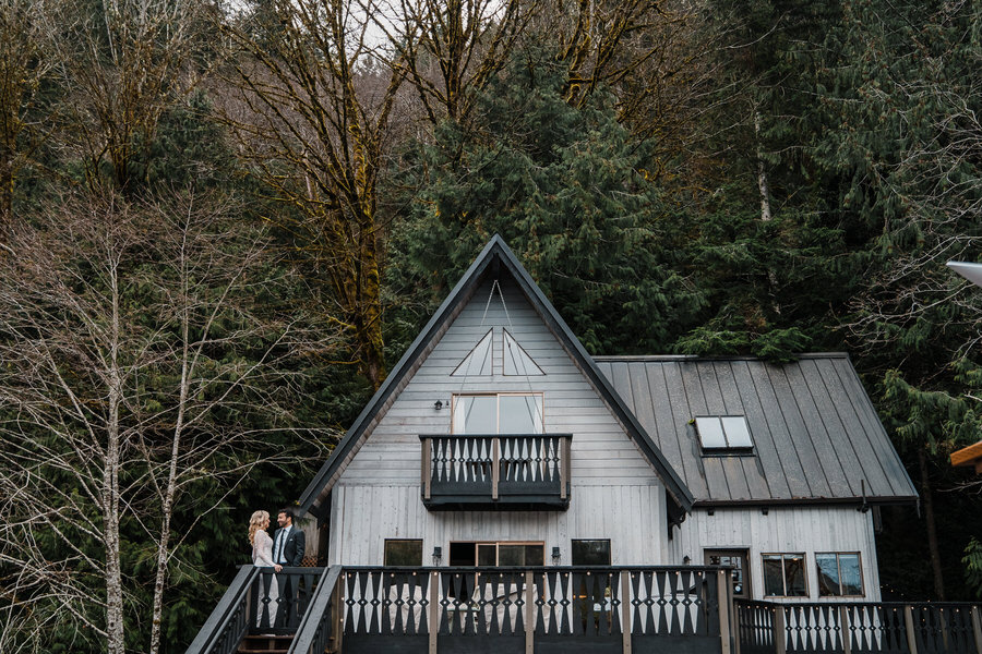 A bride and groom stand on the deck of a lakefront cabin on Lake Sutherland while tall mossy trees surround them