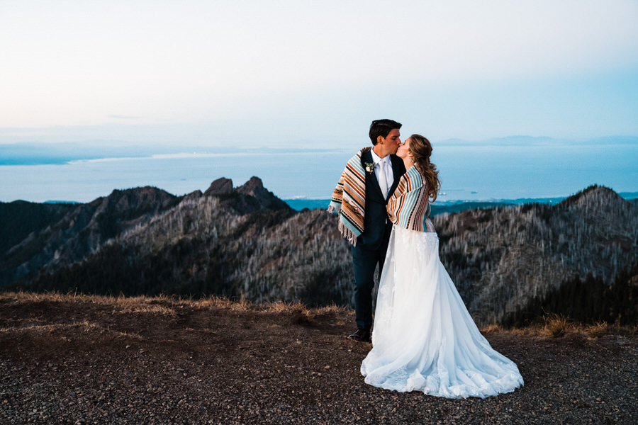 a bride and groom snuggle under a blanket with the Olympic Mountains and Strait of Juan de Fuca in the background at twilight
