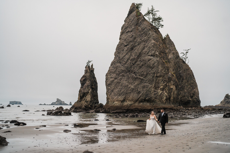 a bride and groom walk hand in hand on rialto beach with dramatic sea stacks in olympic national park