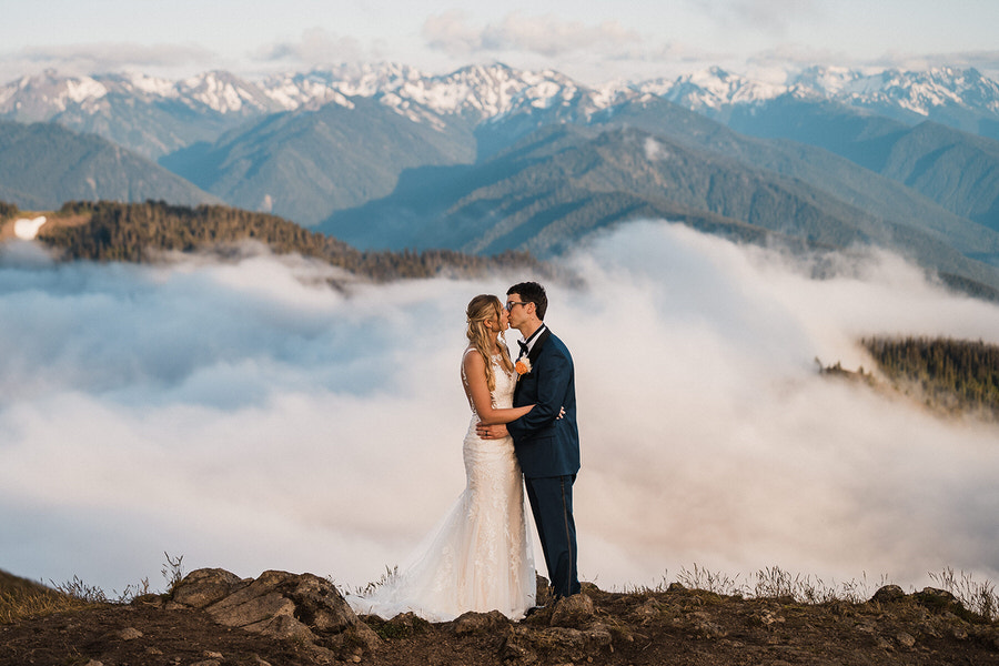 a couple kisses in front of a vast mountain view above the clouds on their wedding day