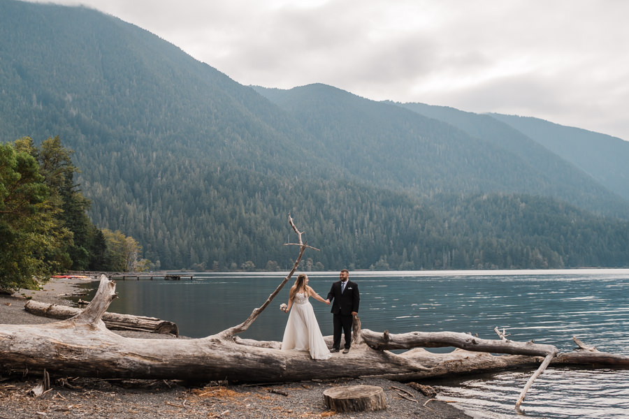 a bride and groom walk across a log on their elopement day at lake crescent in olympic national park